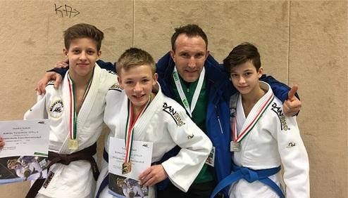Vier Personen in Judoanzügen posieren lachend mit Medaillen und Urkunden vor einer neutralen Wand.