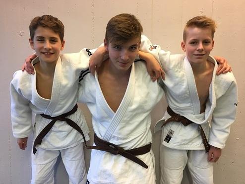 Drei Jungs in Judoanzügen mit braunen Gürteln stehen lächelnd vor einer hellen Wand.