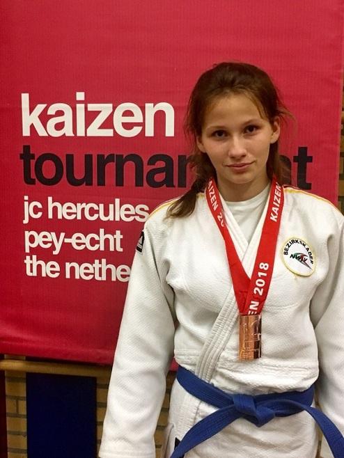 Mädchen in Judoanzug mit bronzener Medaille steht vor rotem Banner des Kaizen-Turniers.