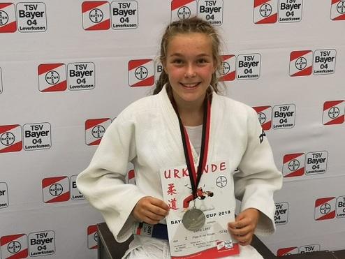 Mädchen in Judogi hält eine Urkunde und Medaille, sitzt vor einer Wand mit Logos des TSV Bayer 04 Leverkusen.