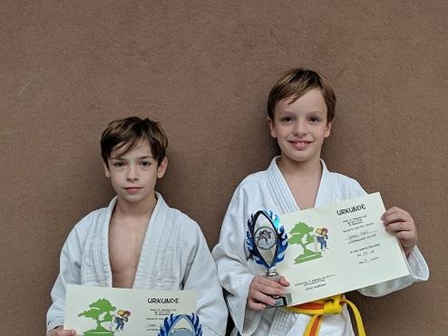 Zwei Kinder in Judo-Anzügen halten Urkunden und einen Pokal vor einer braunen Wand.
