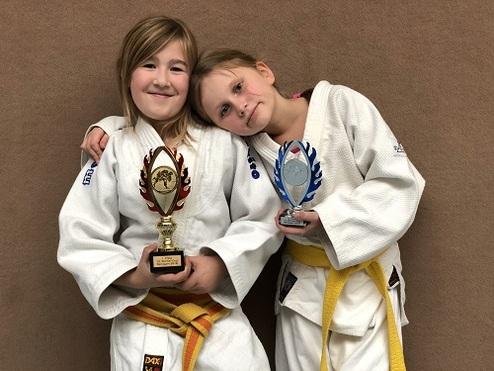 Zwei Mädchen in Judogis halten Trophäen und lächeln vor einem braunen Hintergrund.