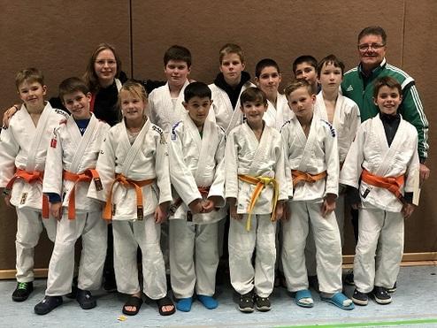 Gruppe von zwölf Kindern in Judogis mit unterschiedlichen Gürtelfarben, umringt von zwei Erwachsenen in der Sporthalle.