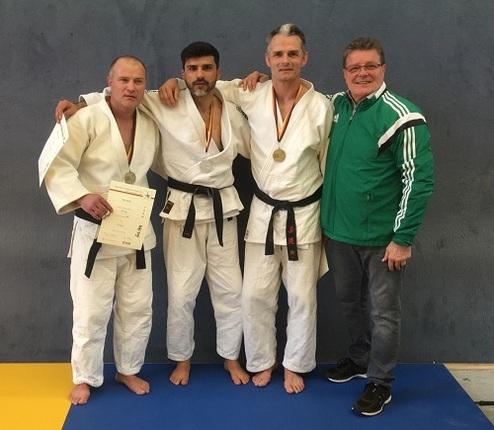 Vier Männer in Judogi stehen auf einer Matte, zwei mit Medaillen, einer hält ein Zertifikat, während der Trainer lächelt.