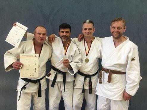 Vier Judokas in Uniform stehen zusammen, halten Urkunden und Medaillen in einer Sporthalle. Hintergrund in Blau.