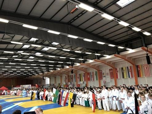 Judo-Wettbewerb: Gruppen von Judokas in weißen Anzügen mit Flaggen auf einer blauen Matte in einer großen Halle.