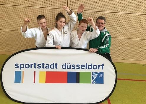 Vier Personen in Judokleidung posieren mit einem Banner der Sportstadt Düsseldorf in einer Sporthalle.
