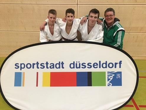 Vier Personen in weißen Judogi stehen lächelnd hinter einem Banner mit der Aufschrift „sportstadt düsseldorf“.