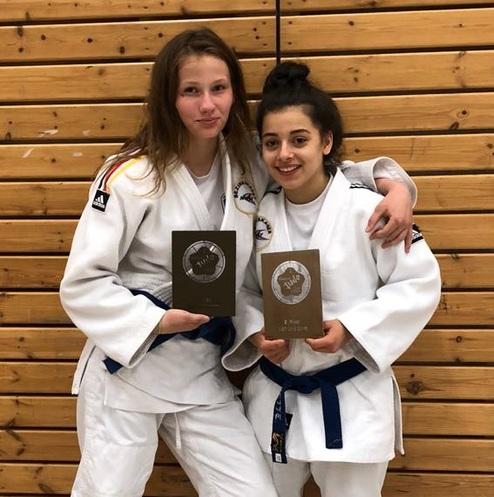 Zwei Judokas in weißen Anzügen halten Pokale in der Hand und posieren lächelnd vor einer Holzwand.