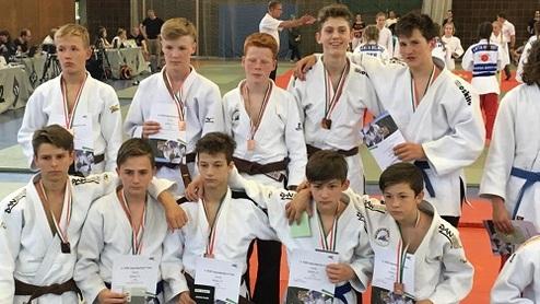 Gruppe von Judokas in weißen Gi mit Medaillen und Urkunden auf einer Wettkampfmatte.