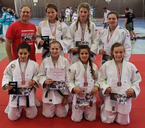 Gruppe von acht Judokas in weißen Gi, steht auf einer Judomatte, alle halten Urkunden und Medaillen.