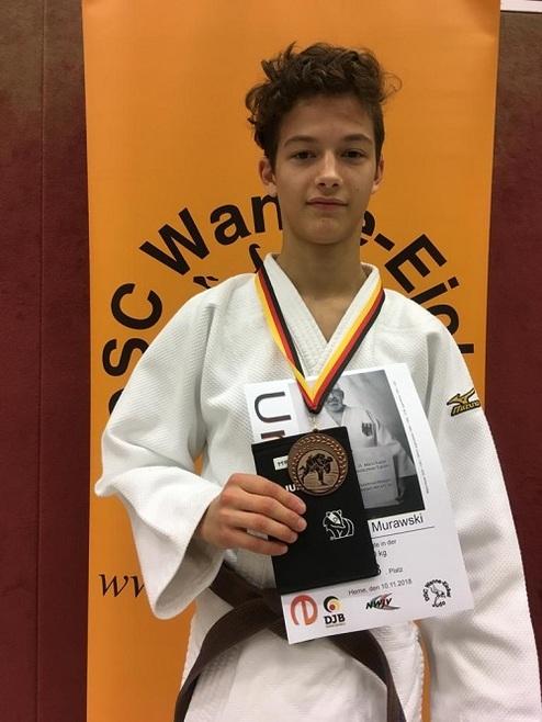 Junger Judoka mit Medaille, Urkunde und gelber Wand im Hintergrund.