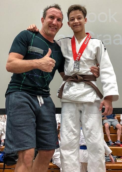 Ein Trainer und ein junger Judoka mit Bronze-Medaille posieren lächelnd nach einem Wettkampf. Judoka trägt einen braunen Gürtel.