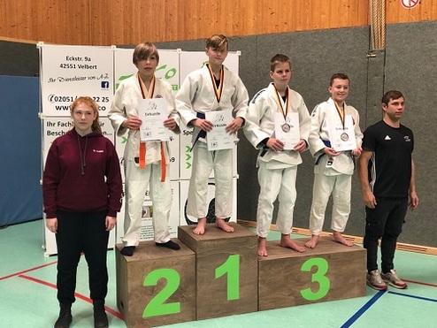 Drei Jugendliche in Judoanzügen stehen auf dem Treppchen und halten Medaillen sowie Urkunden; ein Trainer steht daneben.