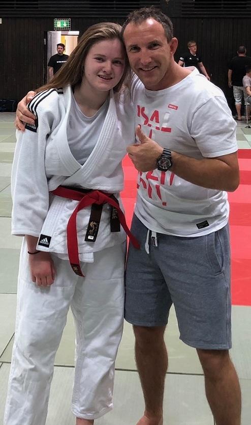 Zwei Personen posieren lächelnd auf einer Judomatte; das Mädchen trägt einen weißen Judogi mit rotem Gürtel.