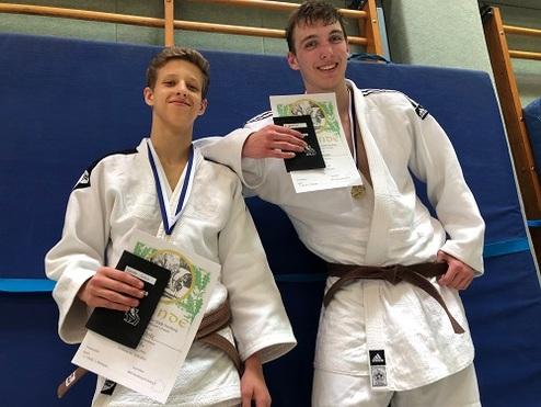 Zwei Judokas in weißen Gi halten ihre Urkunden und Medaillen vor einer blauen Wand.