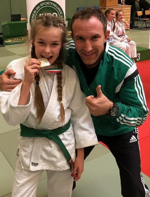 Lächelnde junge Judoka mit Medaille in der Hand, posierend mit Trainer auf einer Judomatte im Hintergrund.