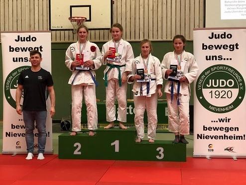 Auf dem Siegerpodest stehen vier Judokas mit Medaillen, neben ihnen ein Trainer. Im Hintergrund sind Banner mit Judosprüchen.