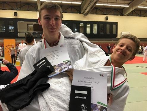 Zwei Judokas in weißen Anzügen halten Urkunden und einen Judo-Pass in einer Sporthalle.