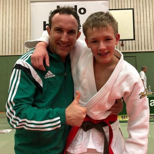 Ein Trainer umarmt einen jungen Judoka, beide lächeln und zeigen Daumen hoch in einer Sporthalle.