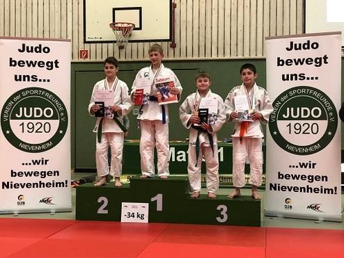 Vier Judoka stehen auf dem Siegerpodium mit Medaillen und Pokalen, umgeben von Werbebannern des Vereins.