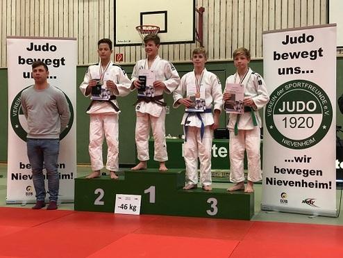 Vier Judoka in weißem und grünem Gi stehen auf dem Treppchen und halten Medaillen und Urkunden in einer Sporthalle.