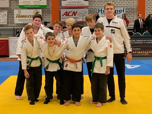 Gruppe junger Judoka in weißen Judokas mit grünen und schwarzen Gürteln auf einer Judomatte, lächelnd und posierend.