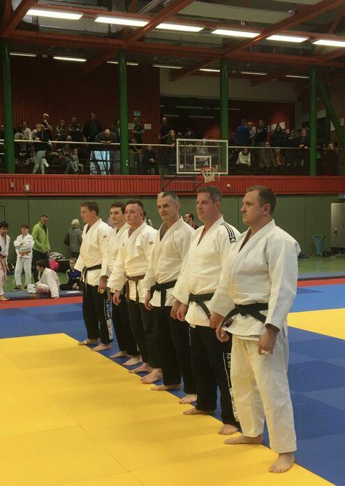 Sechs Männer in Judogis stehen auf einer Mattenfläche, bereit für einen Wettkampf, Publikum im Hintergrund.