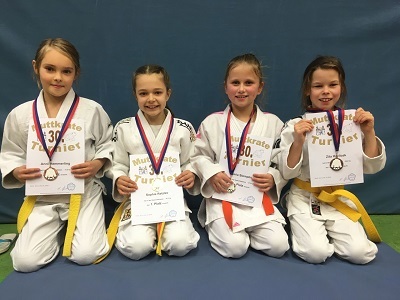 Vier Kinder in Karateanzügen mit Medaillen und Urkunden an einem Tisch stehen lächelnd vor einer blauen Wand.