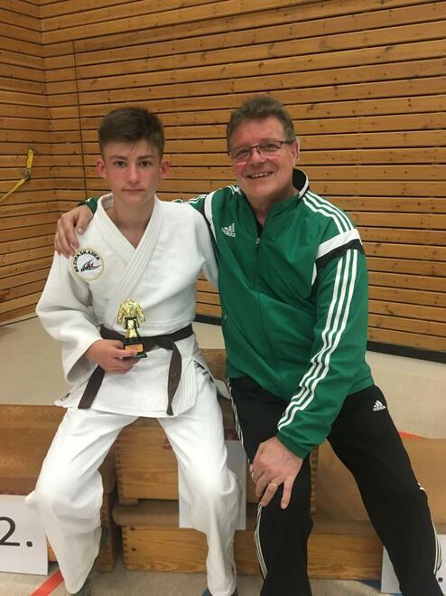 Junge in Judoanzug mit braunem Gürtel hält einen Pokal, sitzt neben einem lächelnden Trainer in sportlicher Kleidung.