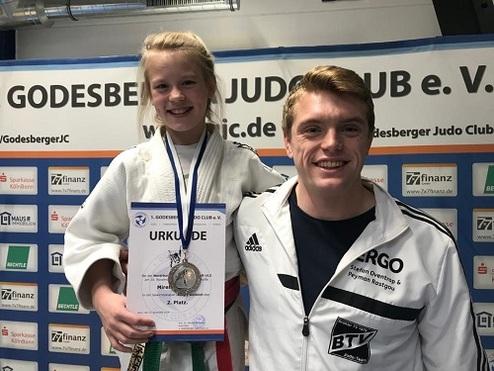 Mädchen in Judog uniform hält Urkunde und Medaille, steht lächelnd neben Trainer vor Godesberger Judo-Club Banner.