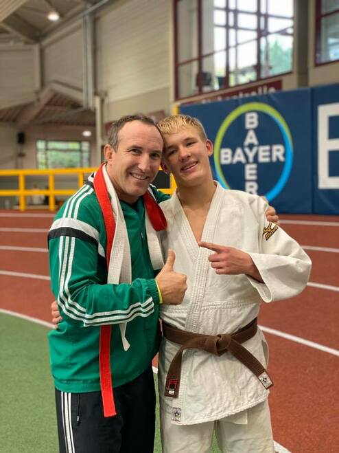 Zwei Männer stehen arm in arm in einer Sporthalle, einer trägt einen Judoanzug mit braunem Gürtel, der andere hat einen grünen Sportpullover an.
