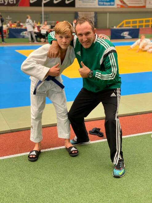 Junge in Judogi und Trainer in Sportkleidung posieren lächelnd auf einer Judomatte in einer Sporthalle.