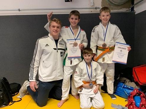 Gruppe von vier Judokas mit Medaillen und Urkunden auf einer Judomatte, ein Trainer kniet vor ihnen.