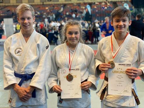 Drei Kinder in Judoanzügen halten Urkunden und Medaillen nach der Bremen Open Veranstaltung. Hintergrund zeigt Sportveranstaltung.