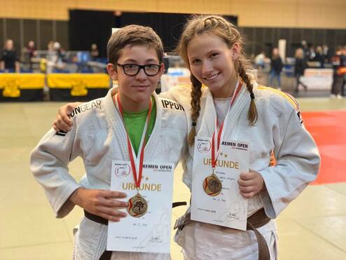 Zwei Jugendliche in Judoanzügen lächeln stolz mit Medaillen und Urkunden nach einem Wettkampf.