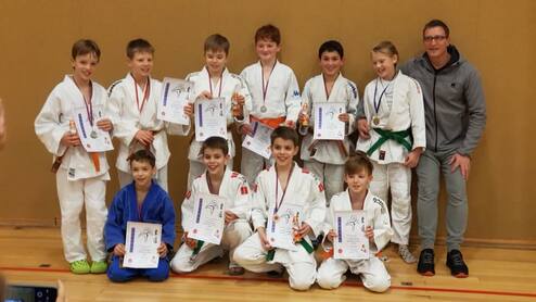 Gruppe von zwölf Kindern in Judoanzügen hält Urkunden und Medaillen, während ein Trainer lächelnd daneben steht.