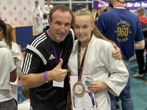 Junge Karateka mit Medaille posiert mit Trainer, beide lächeln vor einer Wettkampfumgebung.