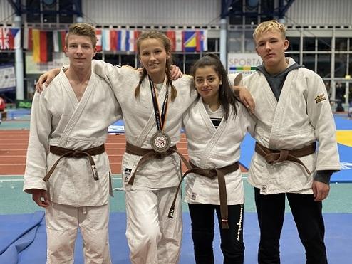 Vier Judosportler in weißem Gi und braunem Gürtel posieren gemeinsam, eine Sportlerin trägt eine Medaille.