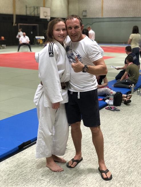 Ein junger Judo-Sportler in weißem Gi umarmt einen Trainer auf einer Judo-Matte in einer Sporthalle.