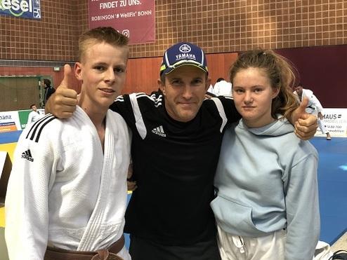 Drei Personen posieren auf einer Judo-Matte, zwei Jugendliche in Judogis und ein Trainer in Sportkleidung.