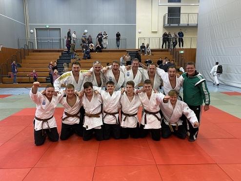 Gruppe von Judoka in weißen Gis posiert lächelnd auf einer roten Judomatte, Zuschauer im Hintergrund.