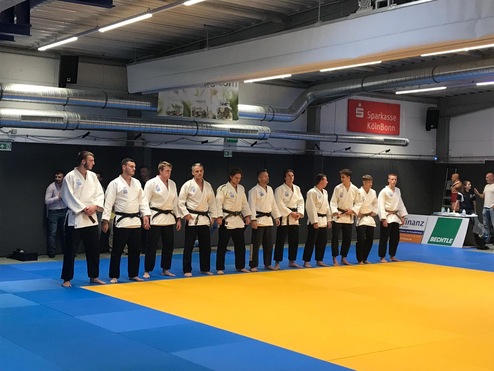 Gruppenszene von Judokas in weißen Gewändern auf einer blau-oranger Matte, bereit für eine Wettkampfvorrunde.