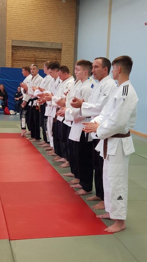 Gruppe von Judoka in weißen Judogis steht auf einer Matte und applaudiert während einer Veranstaltung oder Zeremonie.