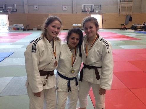 Drei Judokas in weißen Judogis mit Medaillen posieren lächelnd auf einer farbigen Judomatte.