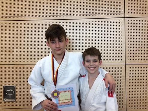 Zwei Kinder in Judoanzügen posieren lächelnd mit einer Urkunde und einer Medaille vor einer Holzpaneelwand.