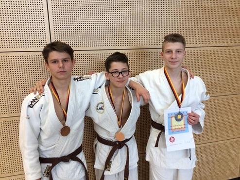 Drei Jugendliche in Judoanzügen stehen zusammen, tragen Medaillen und halten eine Urkunde in der Hand.