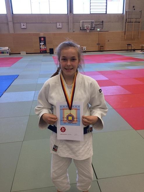Mädchen in Judokleidung hält eine Urkunde mit Medaille in einer Sporthalle auf einer mit Teppichen belegten Matte.