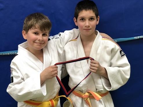 Zwei Jungen in Judoanzügen halten gemeinsam eine Medaille, vor einem blauen Hintergrund mit einem Sportband.