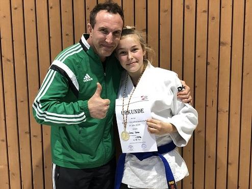 Trainer mit Daumen hoch und Judoka mit Medaille und Urkunde vor Holzpaneelen.